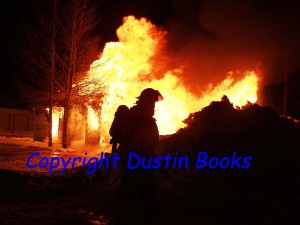 A firefighter is silhouetted against the flames of a fully-involved structure fire.